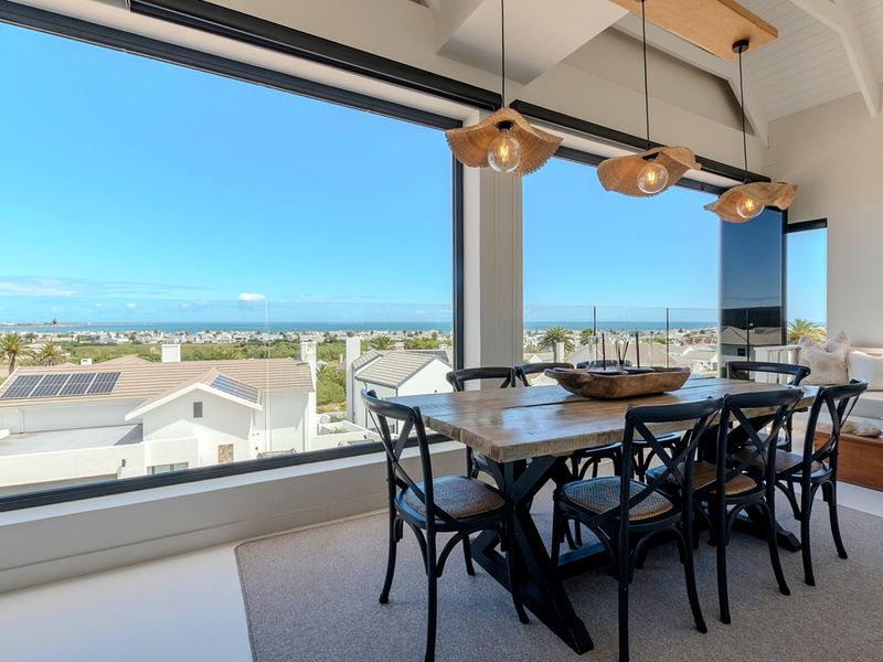 A bright dining area featuring vaulted ceilings with exposed beams and expansive floor-to-ceiling windows offering panoramic coastal and neighborhood views.