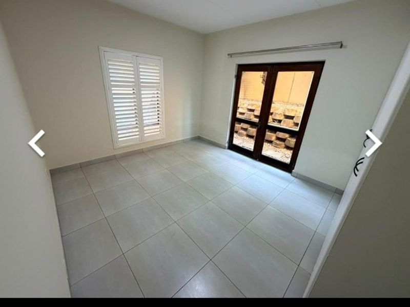 A bright, empty bedroom featuring neutral tiled flooring, plantation shutters, and double glass doors leading to an outdoor area.