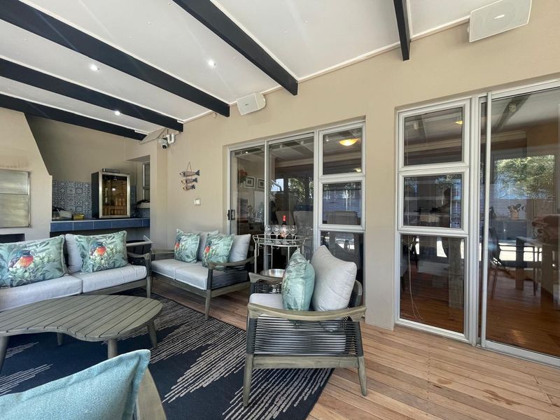Covered patio featuring timber decking, exposed ceiling beams, recessed lighting, and a built-in braai area with tiled counter, seamlessly connected via sliding glass doors.