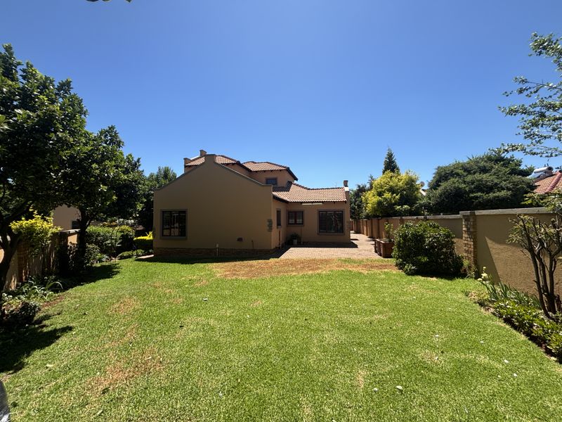 A view of the rear exterior of the house featuring a well-maintained lawn, paved patio area, and surrounding boundary walls.