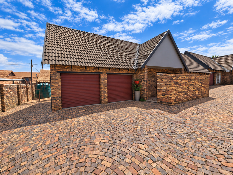 Front exterior view of a townhouse featuring a double garage with red roller doors, brick facade, and a well-maintained circular pattern paved driveway.