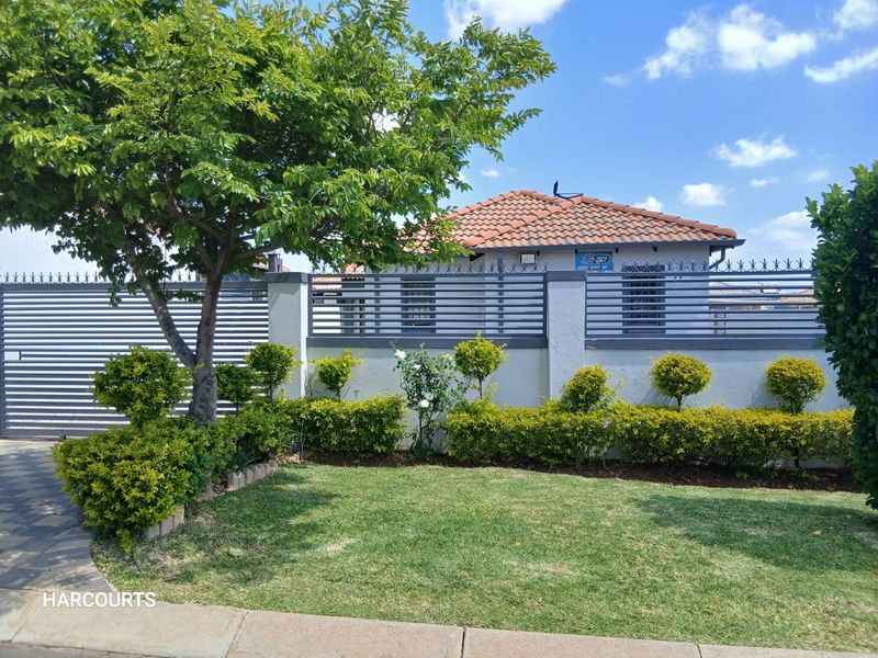 Front view of the property featuring a well-maintained garden, paved driveway, and secure perimeter wall with modern metal slat fencing.