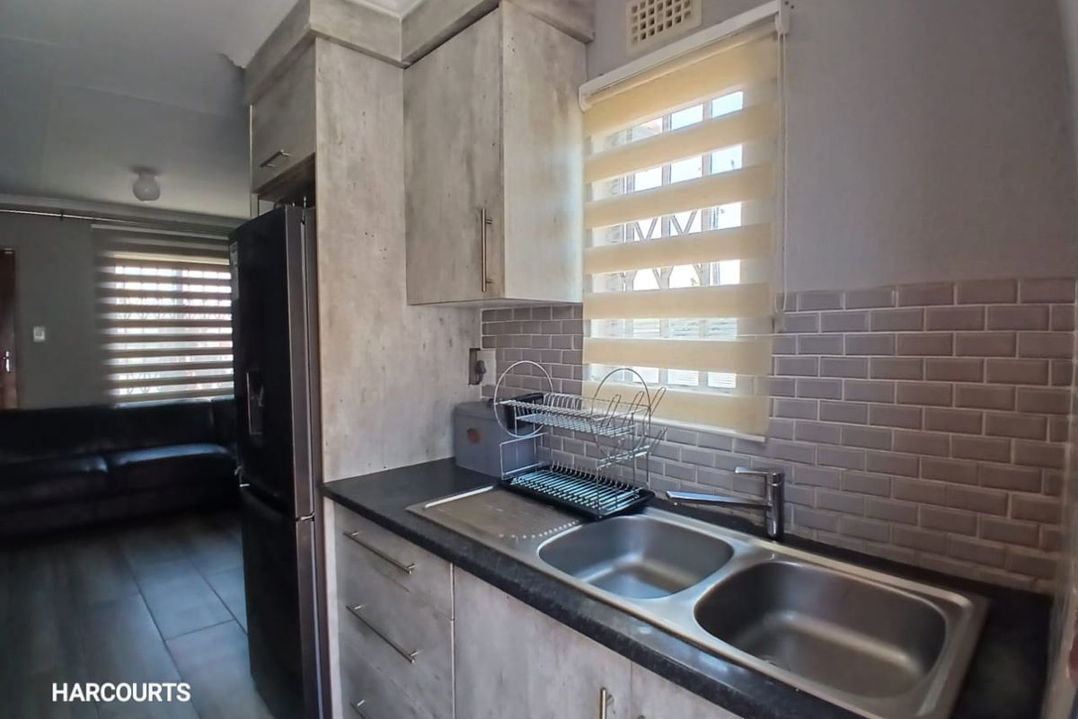 A compact kitchen featuring modern grey-toned cabinetry, subway tile backsplash, and a double bowl stainless steel sink with a view into the adjacent living area.