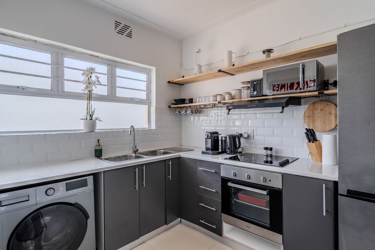 Modern kitchen featuring dark cabinetry, subway tile backsplash, open timber shelving, integrated oven, and cooktop with a convenient laundry connection.