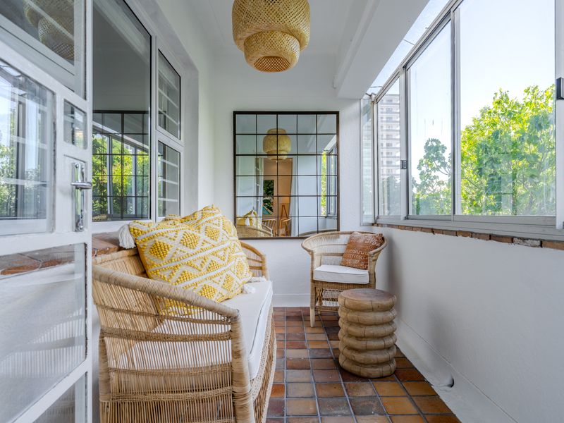 A bright, enclosed balcony featuring terracotta tile flooring, large sliding glass windows, and a decorative wall mirror, offering a pleasant outdoor seating area.