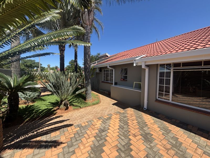 A well-maintained rear exterior view featuring a paved patio area, landscaped garden with mature palms, and a tiled roof structure.