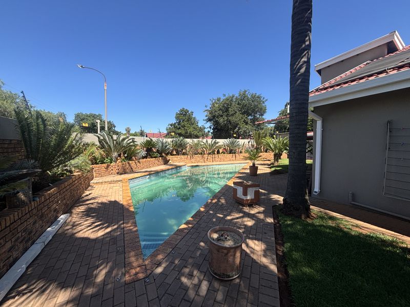 A well-maintained outdoor pool area featuring a triangular swimming pool, brick paved surround, and landscaped retaining walls with perimeter security fencing.