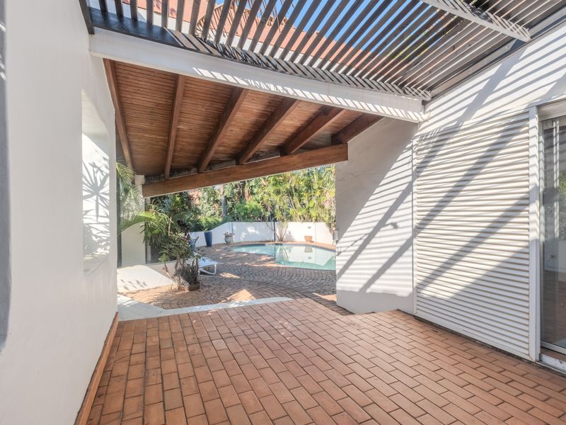 Covered patio area featuring timber pergola and exposed beams, overlooking a paved pool area with direct access to the interior via sliding glass doors.