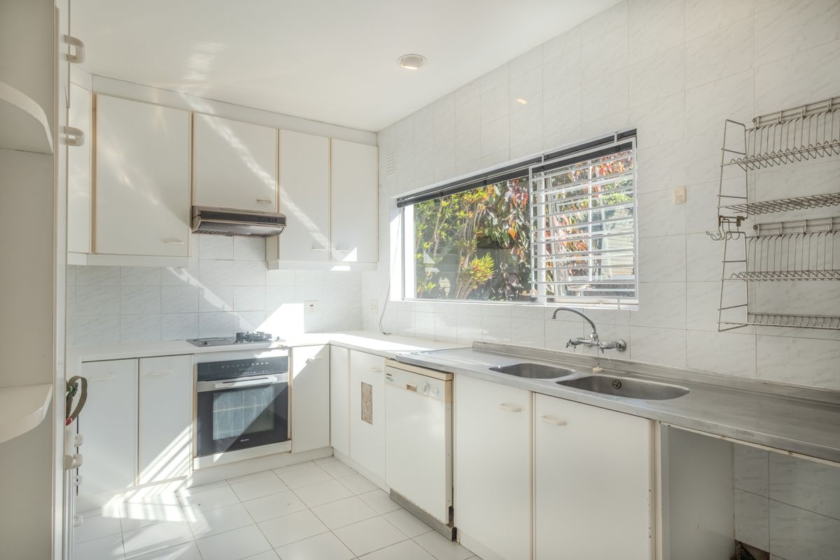 A functional kitchen featuring white cabinetry, stainless steel sink, built-in oven, hob, and extractor fan with tiled splashbacks and flooring.