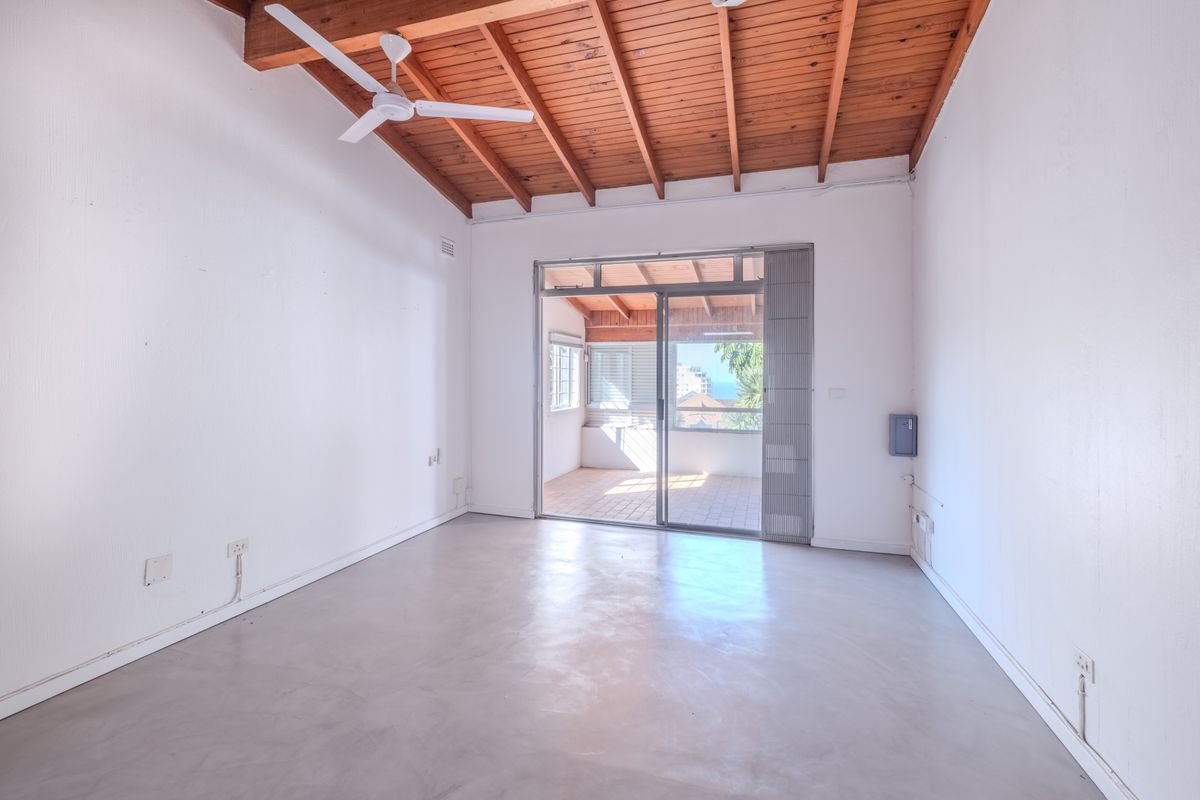 An empty living room featuring exposed timber beam ceilings, concrete flooring, and a sliding glass door leading to an outdoor patio area.