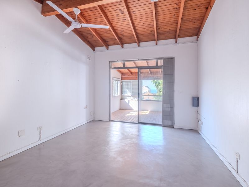 An empty living room featuring exposed timber beam ceilings, concrete flooring, and a sliding glass door leading to an outdoor patio area.