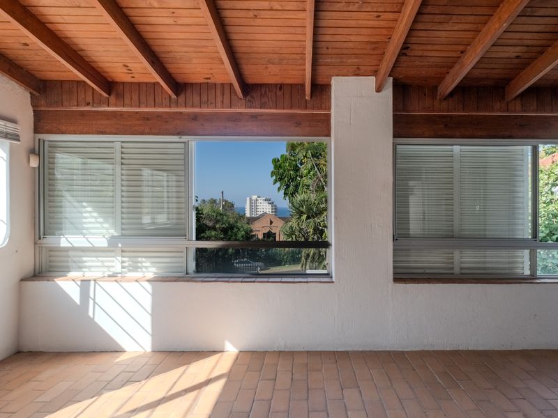 A bright, enclosed patio area featuring a rustic exposed timber ceiling, terracotta tiled flooring, and large windows offering pleasant neighbourhood and distant sea views.