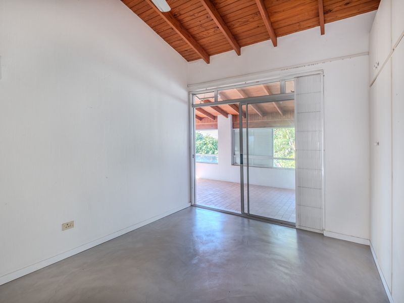 Bright bedroom featuring polished concrete floors, exposed timber beam ceiling, built-in cupboards, and sliding glass doors opening onto a covered patio.