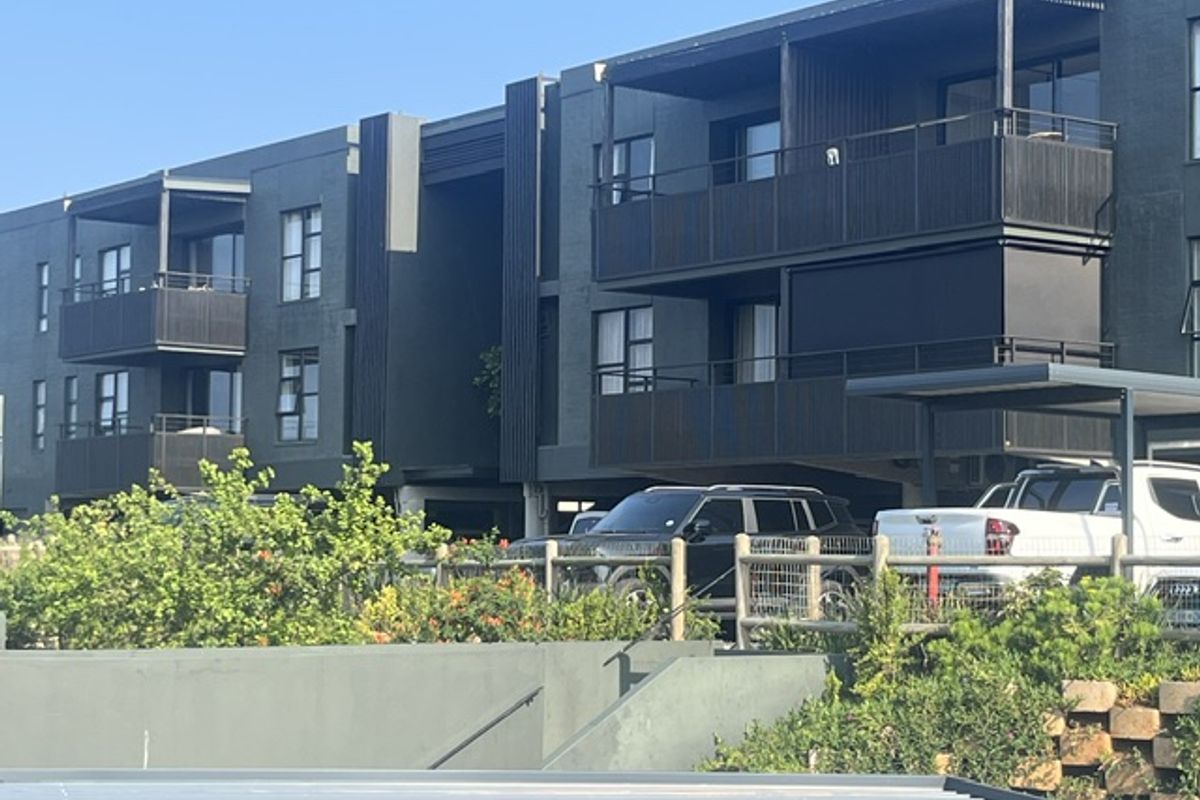 A modern apartment building exterior featuring dark brick cladding, private balconies with metal railings, and an adjacent carport area.