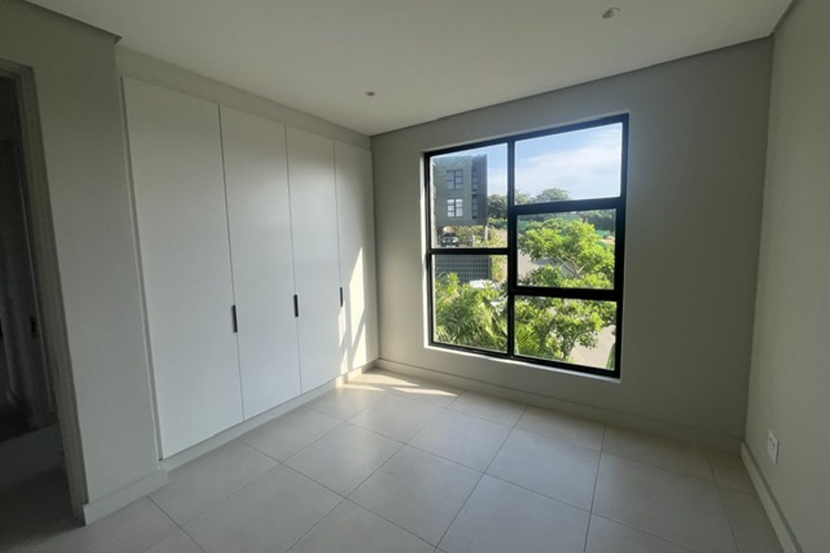 A bright, modern bedroom featuring neutral tiled flooring, ample built-in cupboard space, and large windows providing natural light and pleasant outdoor views.
