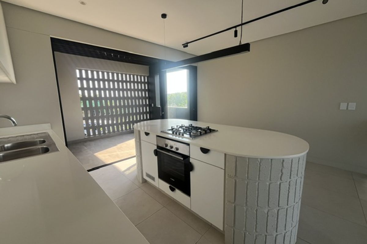 Modern kitchen featuring a central island with built-in oven and gas hob, stone countertops, and sleek track lighting, opening onto a covered balcony.