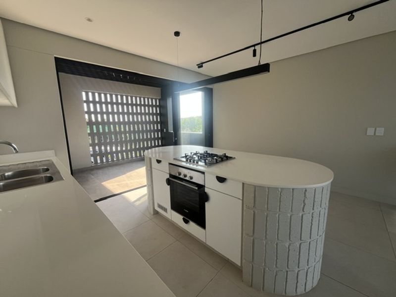 Modern kitchen featuring a central island with built-in oven and gas hob, stone countertops, and sleek track lighting, opening onto a covered balcony.