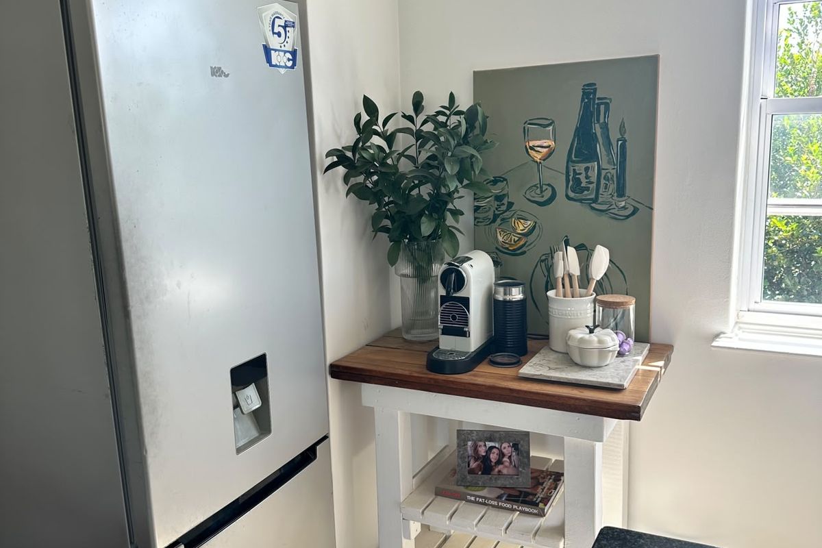 A corner of a kitchen featuring a refrigerator and a small wooden side table positioned near a window.