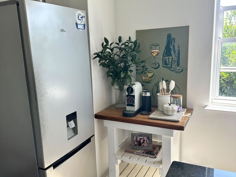 A corner of a kitchen featuring a refrigerator and a small wooden side table positioned near a window.