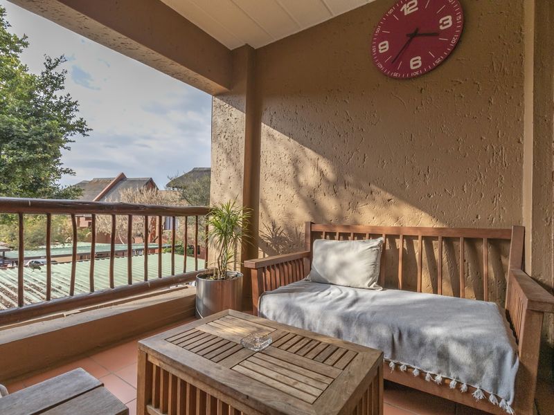 A covered balcony featuring tiled flooring, a wooden balustrade, and textured wall finishes, offering an outdoor space with a view of the surrounding complex.