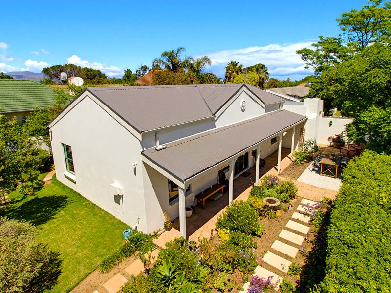 Aerial view of the rear elevation featuring a well-maintained garden, paved pathway, and a covered patio area with a corrugated metal roof.