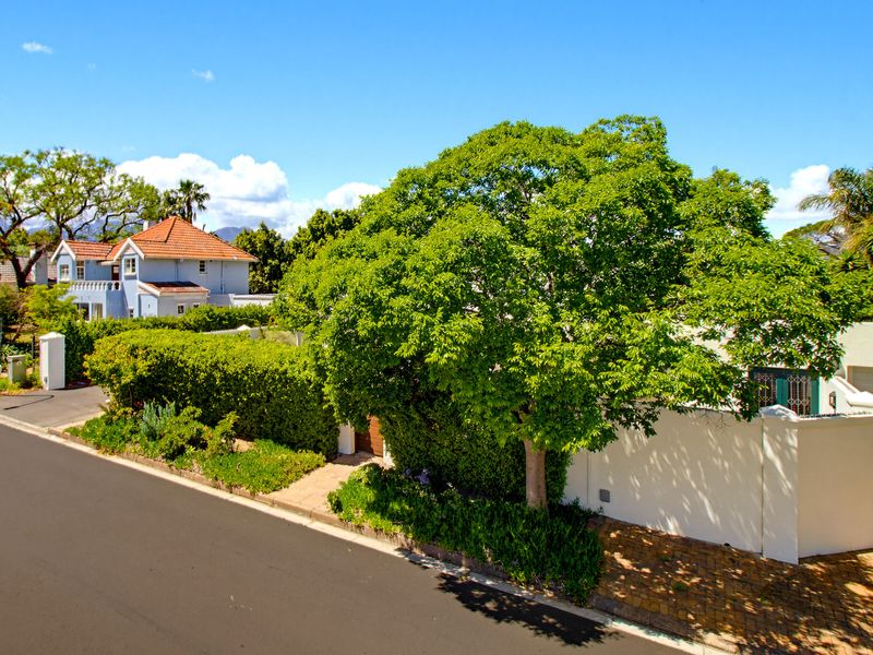 A street-level view of the property featuring a paved driveway, mature landscaping, and a secure boundary wall with a garage entrance.