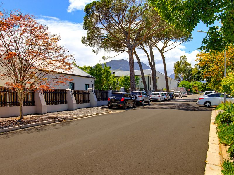 A view of a quiet residential street featuring paved roads, mature trees, and well-maintained boundary walls with mountain views in the background.