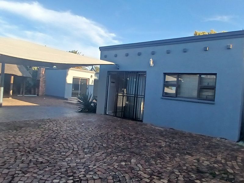Rear exterior view featuring a paved driveway, a large shade sail structure, and a modern blue-painted building with security gates and aluminium windows.