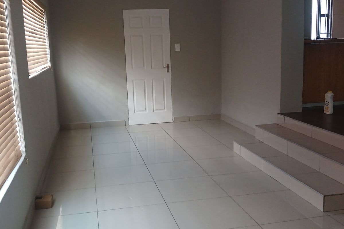 An empty living area featuring neutral tiled flooring, venetian blinds, and a raised platform leading to an adjacent space.