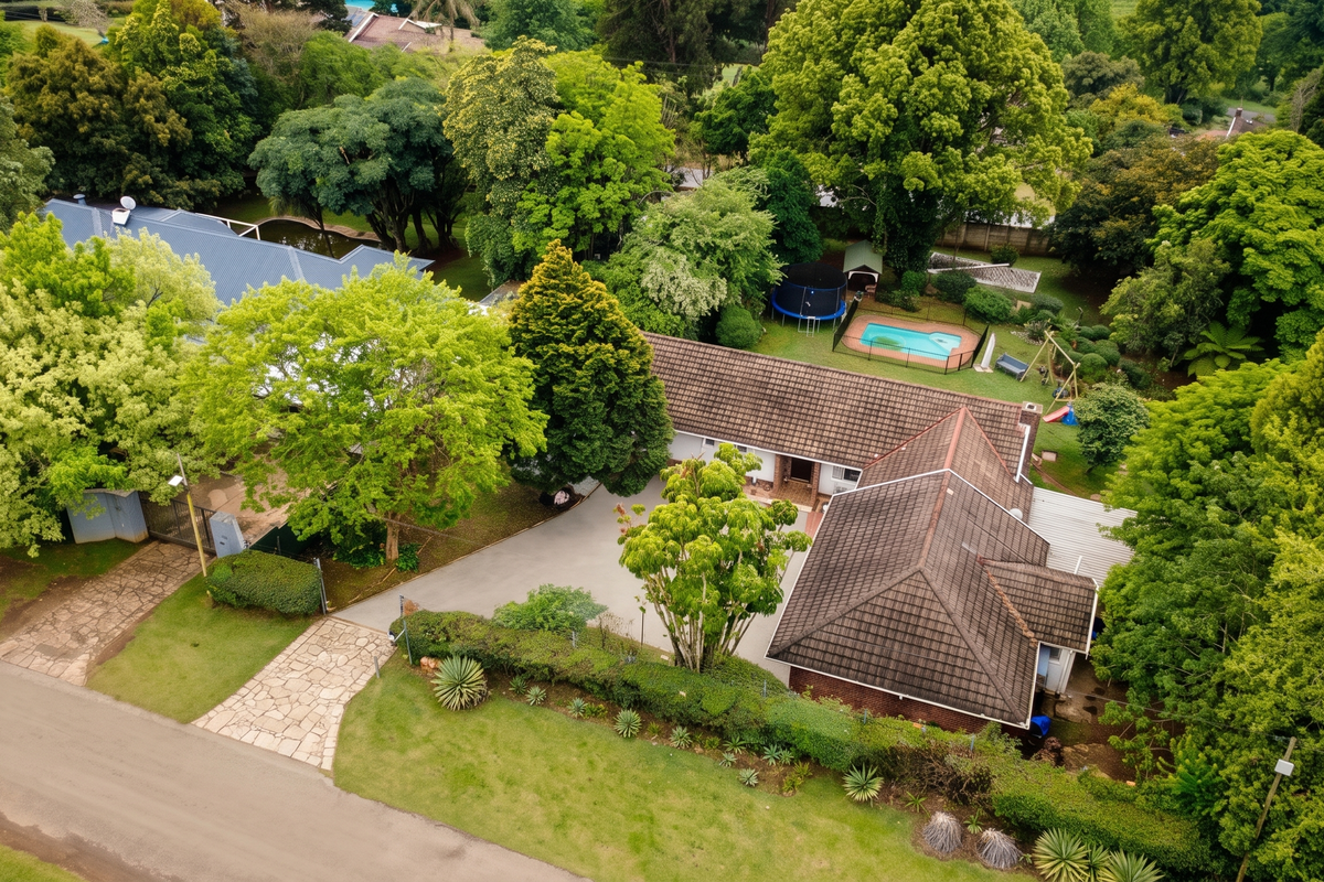 Aerial view of a residential property featuring a spacious garden, swimming pool, tiled roof house, and a paved driveway surrounded by mature trees.