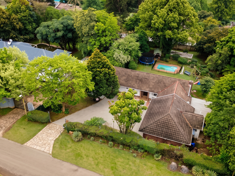Aerial view of a residential property featuring a spacious garden, swimming pool, tiled roof house, and a paved driveway surrounded by mature trees.