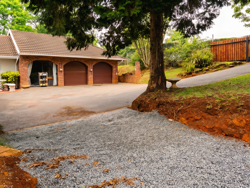 Front exterior view featuring a double garage, asphalt driveway, and established trees, with an unfinished gravel area and exposed soil embankment.