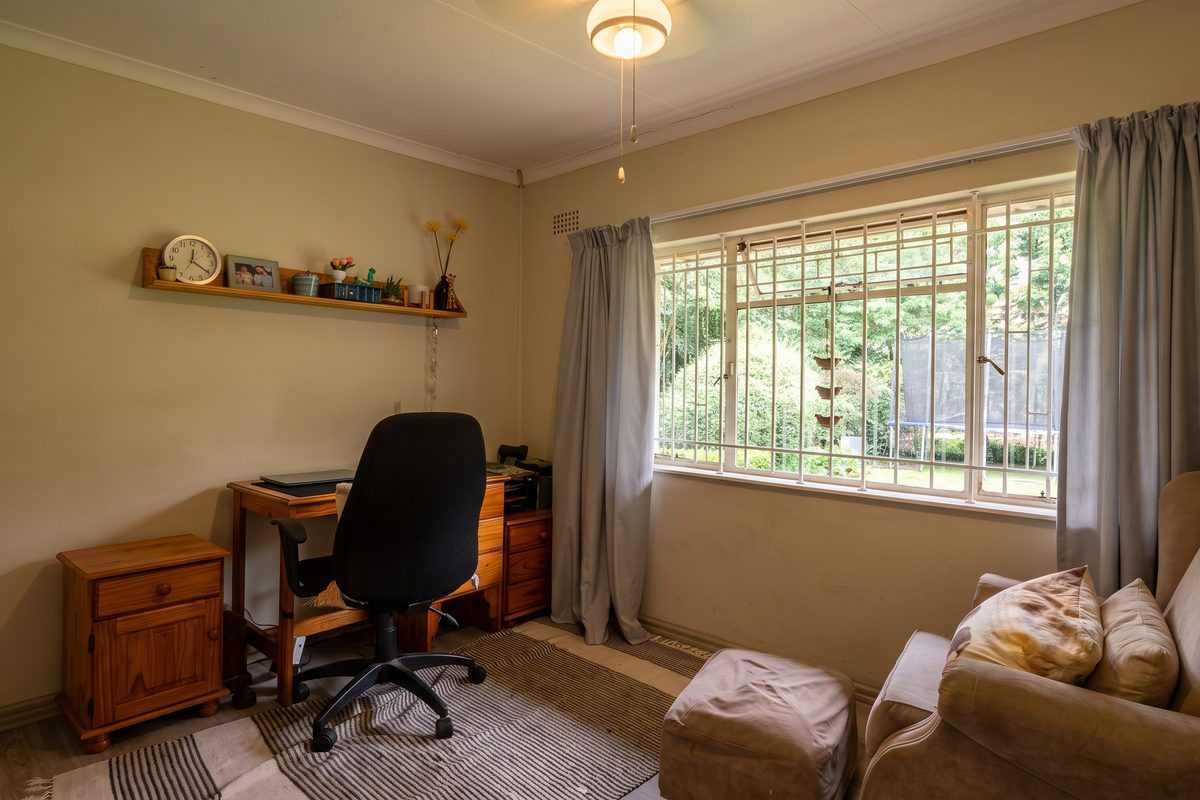 A functional bedroom or home office space featuring security bars on the window and built-in wall shelving, offering a view of the garden.