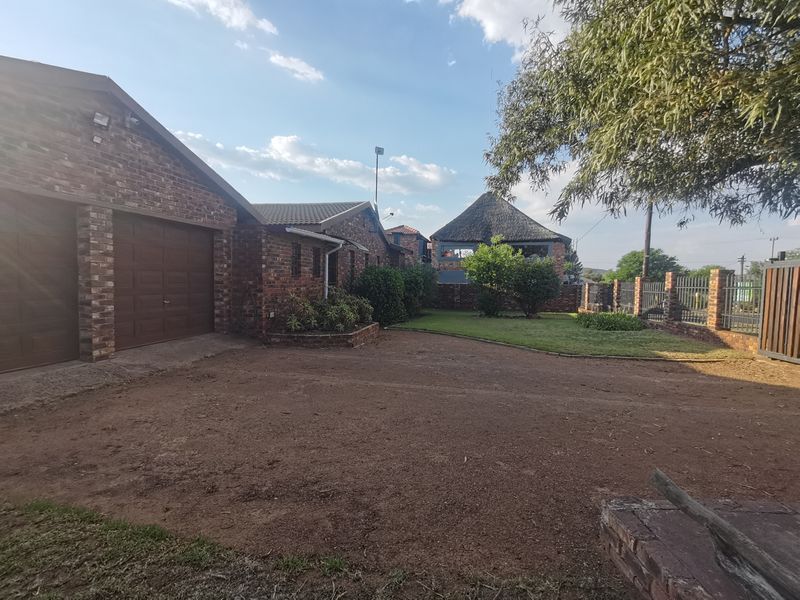 Front view of a face brick house featuring a double garage, paved driveway area, and a landscaped garden with a thatched roof structure in the background.