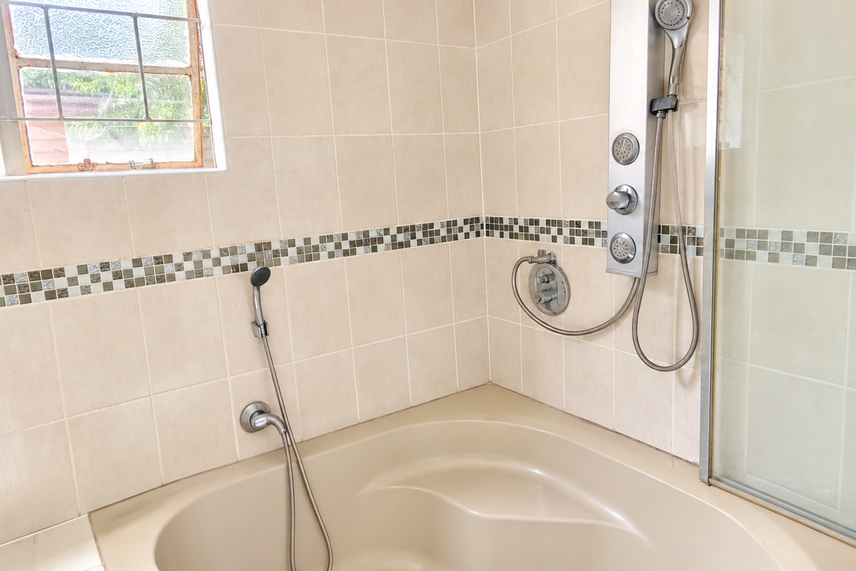 A bathroom featuring a built-in bathtub, a modern shower panel with handheld attachment, and neutral wall tiling with a decorative mosaic border.