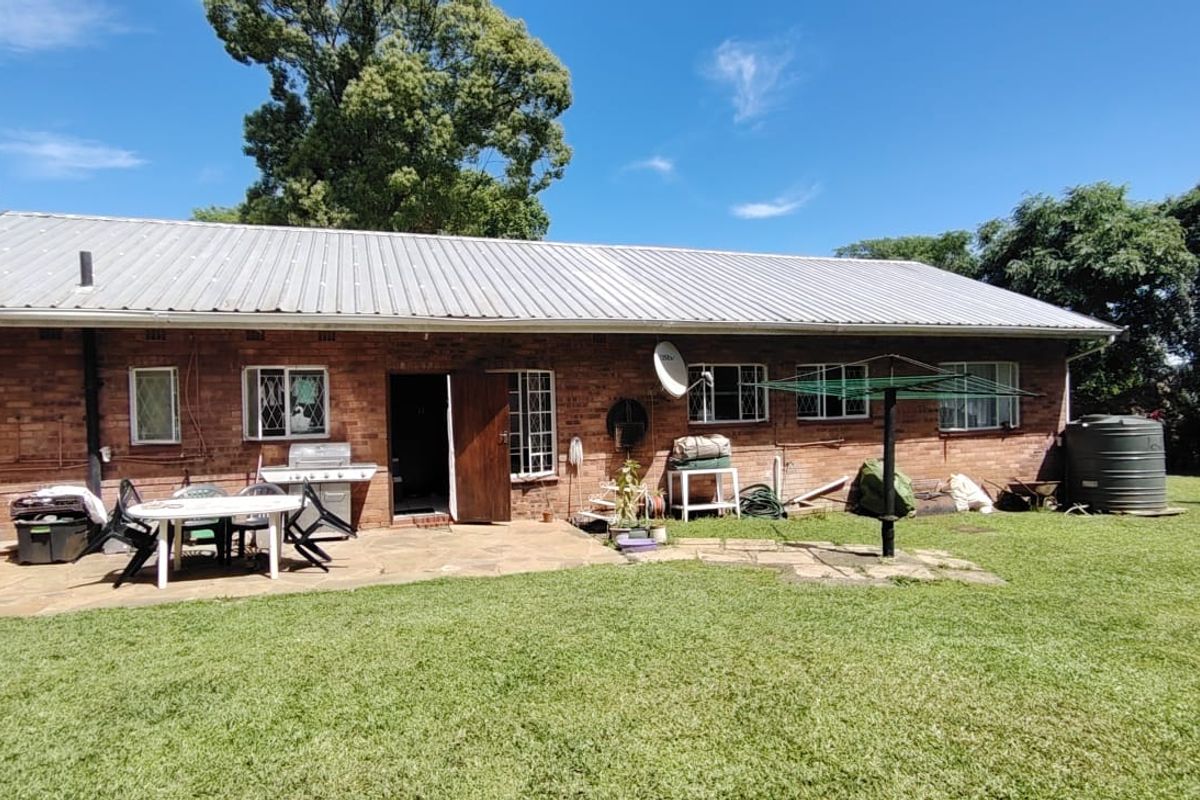 Rear view of a brick house featuring a stone patio, corrugated metal roof, and a large garden area with a water storage tank.