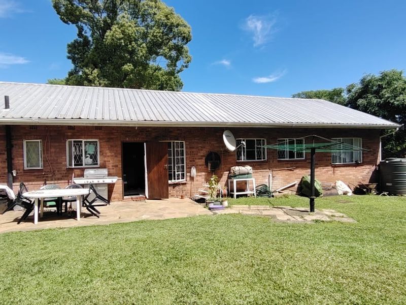 Rear view of a brick house featuring a stone patio, corrugated metal roof, and a large garden area with a water storage tank.