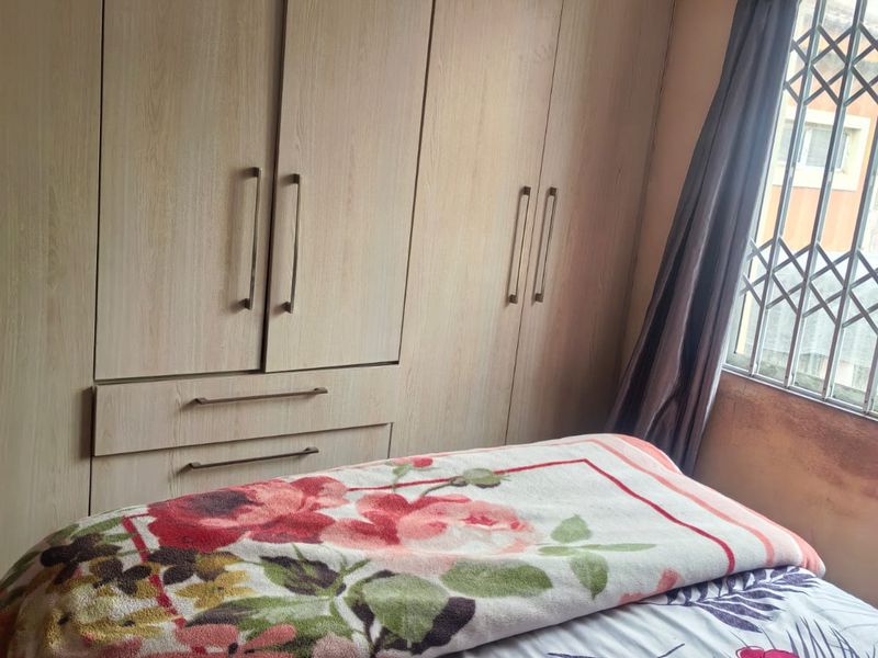 A bedroom featuring built-in wooden cupboards and a window with a security gate. The room shows signs of wear on the walls.