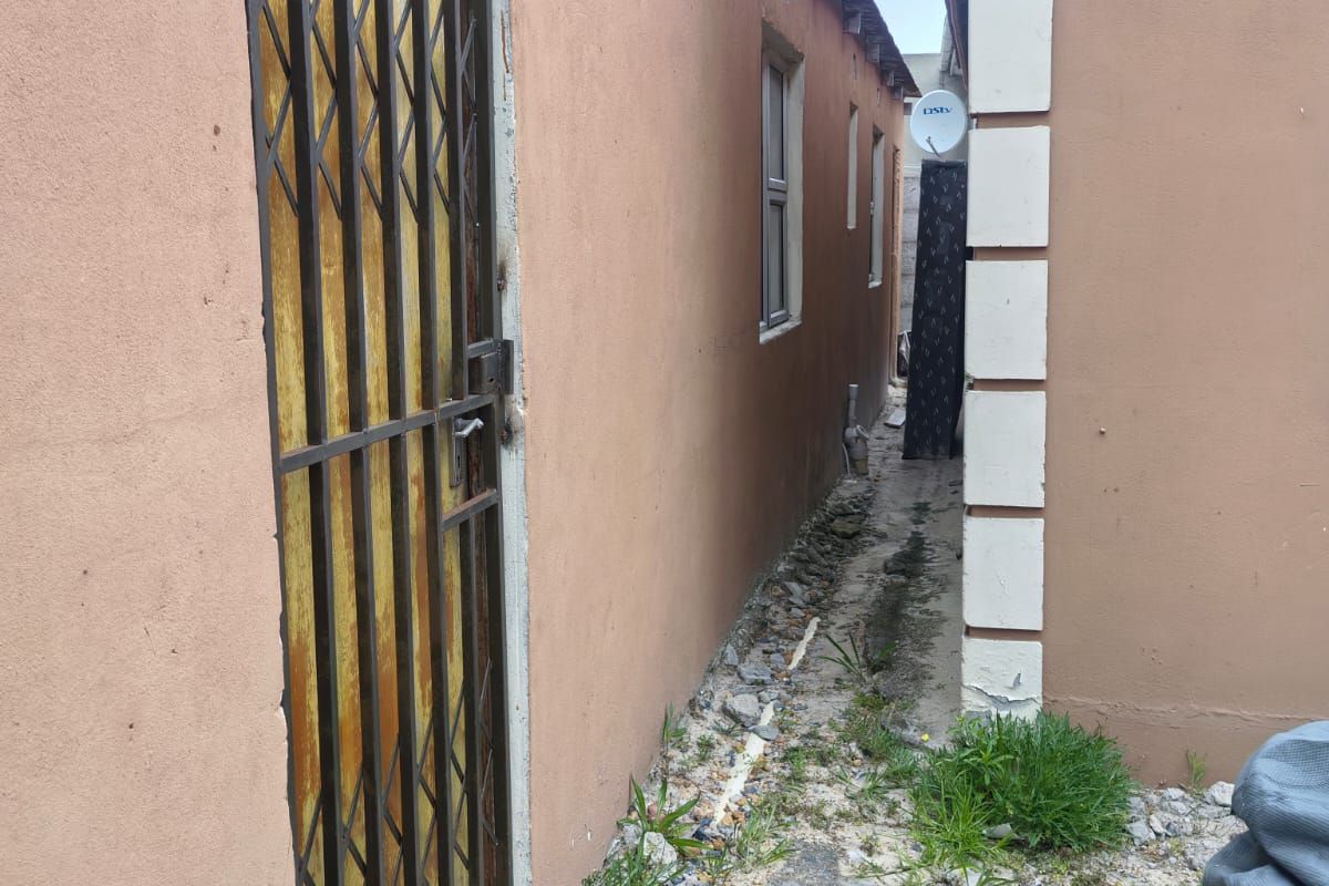 A narrow side passage between two structures featuring a security gate, concrete pathway, and exterior wall finishes requiring maintenance.