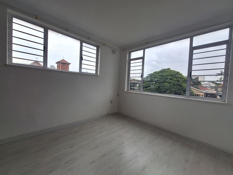 A bright, empty bedroom featuring light-toned laminate flooring and large windows with security bars, offering a clean and neutral space ready for personalization.