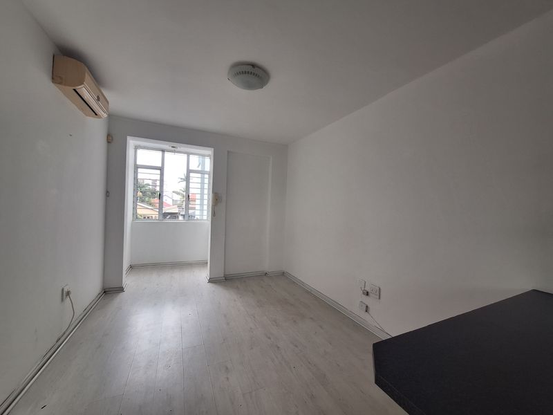 An empty bedroom featuring light-toned laminate flooring, a wall-mounted air conditioning unit, and a window with security bars.