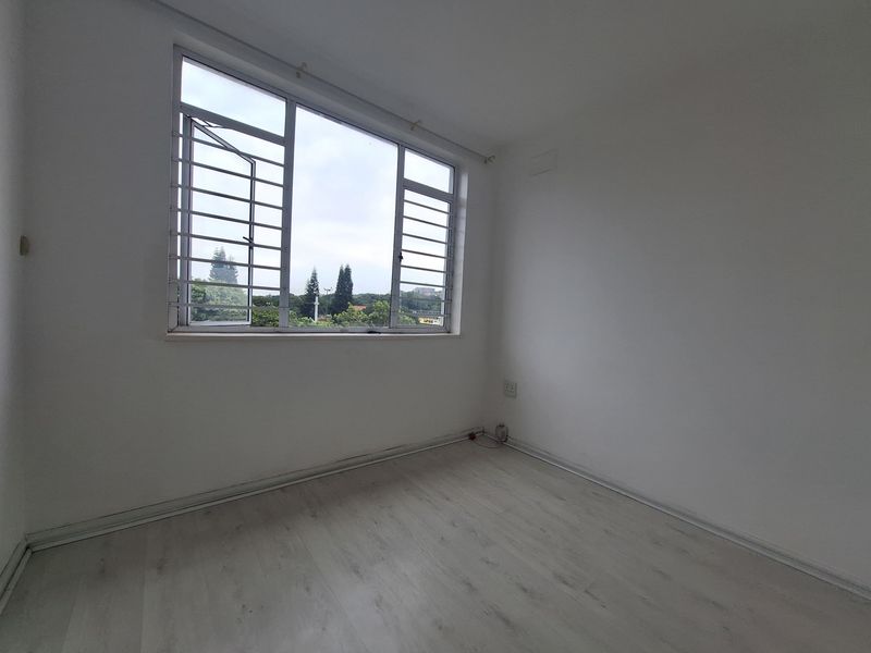 A bright, empty bedroom featuring light-toned laminate flooring, white walls, and a large window fitted with metal security bars.