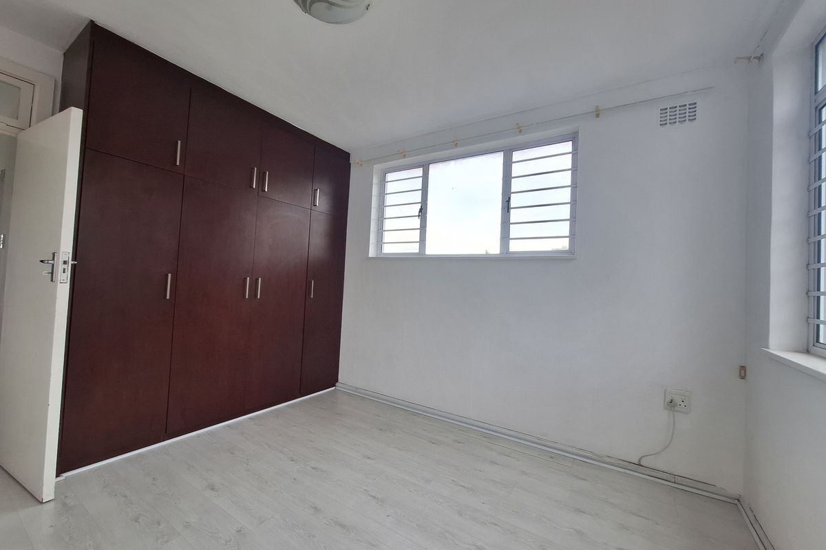 A bright, empty bedroom featuring light-toned laminate flooring, white walls, and dark wood built-in cupboards with secure window burglar bars.