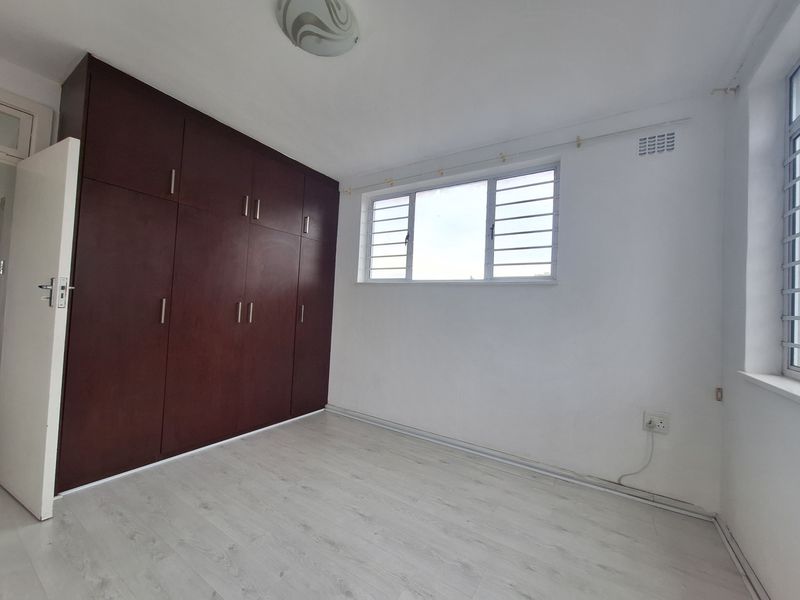 A bright, empty bedroom featuring light-toned laminate flooring, white walls, and dark wood built-in cupboards with secure window burglar bars.