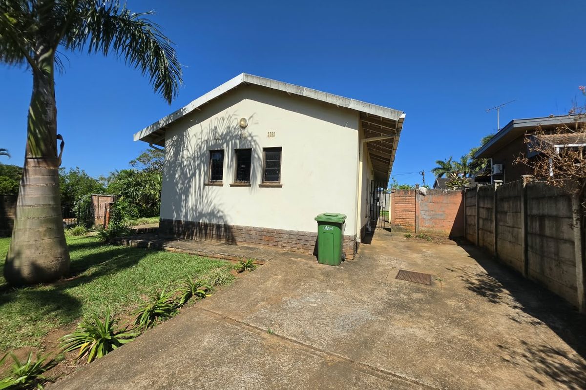 Rear view of the property featuring a concrete driveway, plastered exterior walls, and a brick boundary wall set within a garden area.