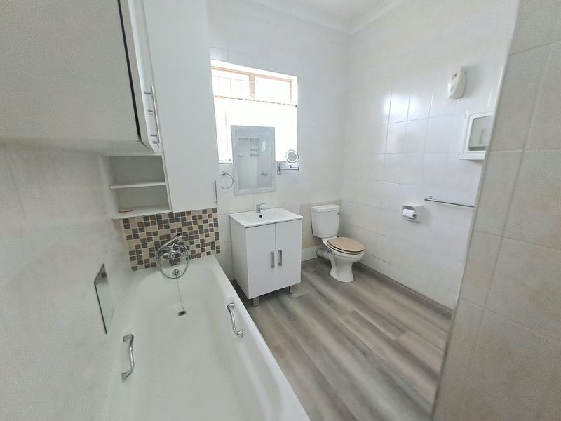A clean and functional bathroom featuring a built-in bathtub, vanity cabinet with basin, and toilet, finished with neutral wall tiles and modern laminate flooring.