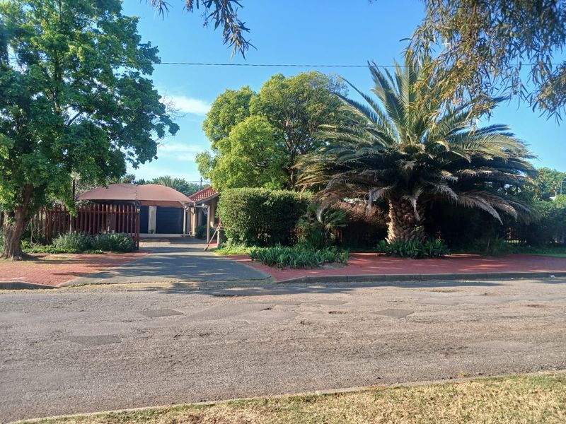 Front view of the property featuring a paved driveway, carport, and established garden with mature trees and palm.