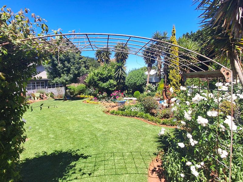 A well-maintained garden featuring a lush green lawn, established flower beds, mature trees, and a decorative metal garden arch.