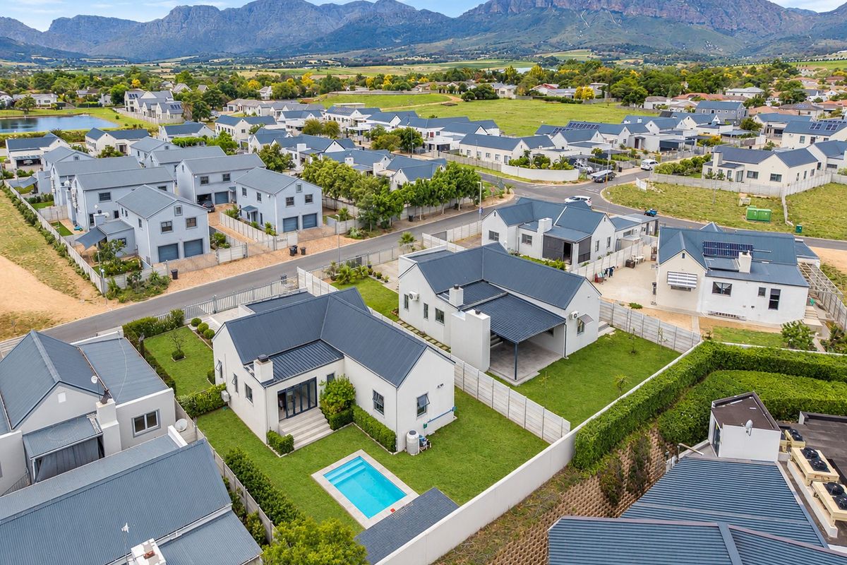 Aerial view of a residential estate featuring detached houses with metal roofs, private swimming pools, and well-maintained gardens against a mountain backdrop.