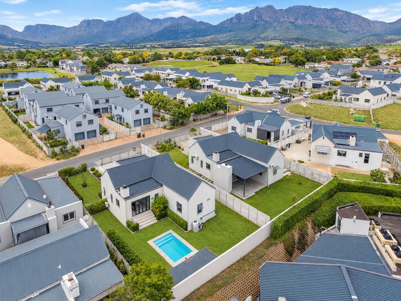 Aerial view of a residential estate featuring detached houses with metal roofs, private swimming pools, and well-maintained gardens against a mountain backdrop.